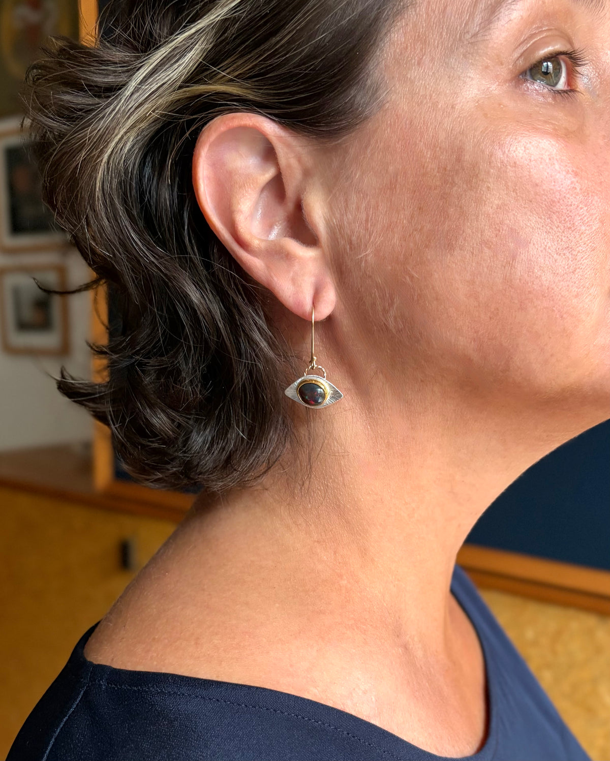 A close-up side profile of a person wearing a dangling, silver, eye-shaped earring.The center stone is a dark gem with flashes of green and red in a gold bezel.