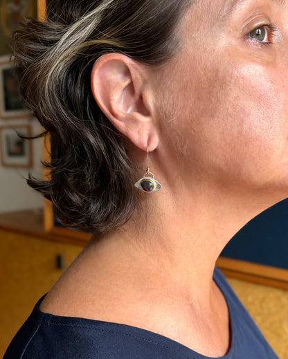 A close-up side profile of a person wearing a dangling, silver, eye-shaped earring.The center stone is a dark gem with flashes of green and red in a gold bezel.