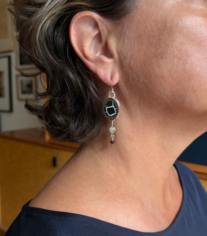 A close-up of a person's ear and neck wearing a dangling earring. The earring features a round black and brown stone with white veining, set in silver, and with small dangling quartz and garnet.