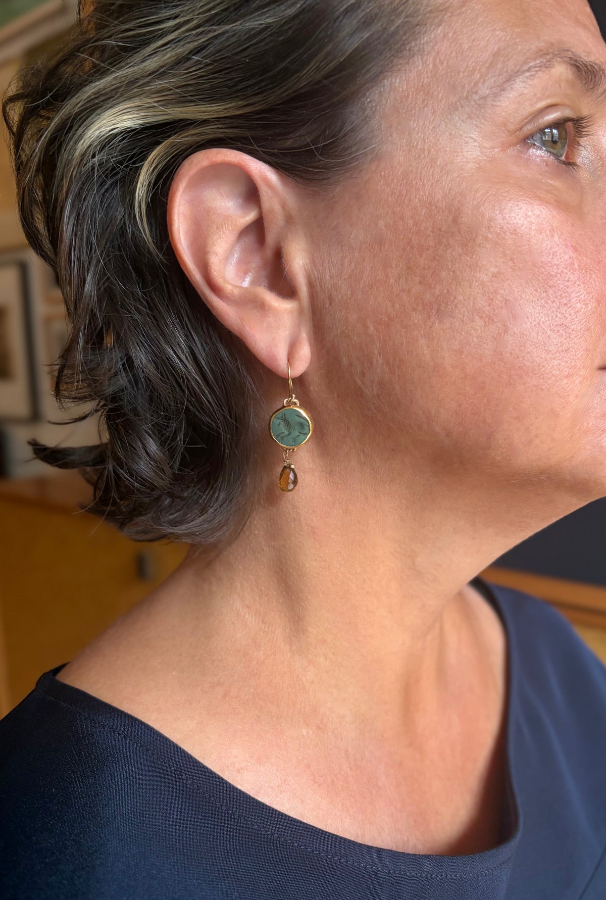 A close-up side profile of a woman wearing a gold dangle earring with a round green stone engraved with the figure of an animal. A small amber drop hangs beneath the central stone. 