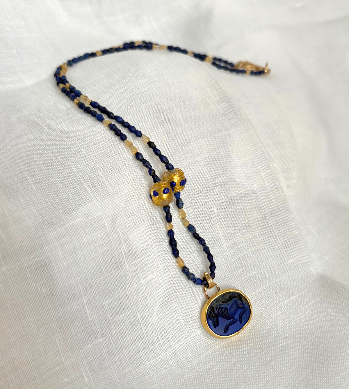 A gold and dark blue beaded necklace with an oval, deep blue lapis, carved with an image of a horse. It is displayed on a textured white fabric background.