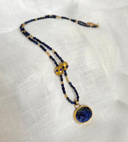 A gold and dark blue beaded necklace with an oval, deep blue lapis, carved with an image of a horse. It is displayed on a textured white fabric background.