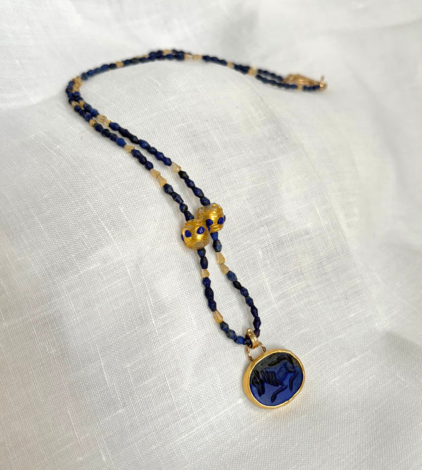 A gold and dark blue beaded necklace with an oval, deep blue lapis, carved with an image of a horse. It is displayed on a textured white fabric background.