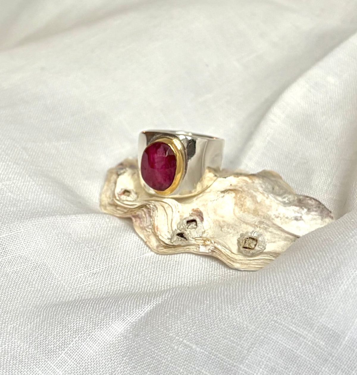 A silver ring with a round, polished deep red rost-cut ruby in a gold bezel setting. It is displayed on an oyster shell on a white fabric background.