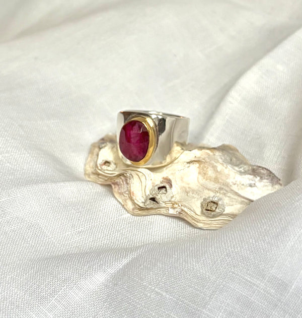 A silver ring with a round, polished deep red rost-cut ruby in a gold bezel setting. It is displayed on an oyster shell on a white fabric background.