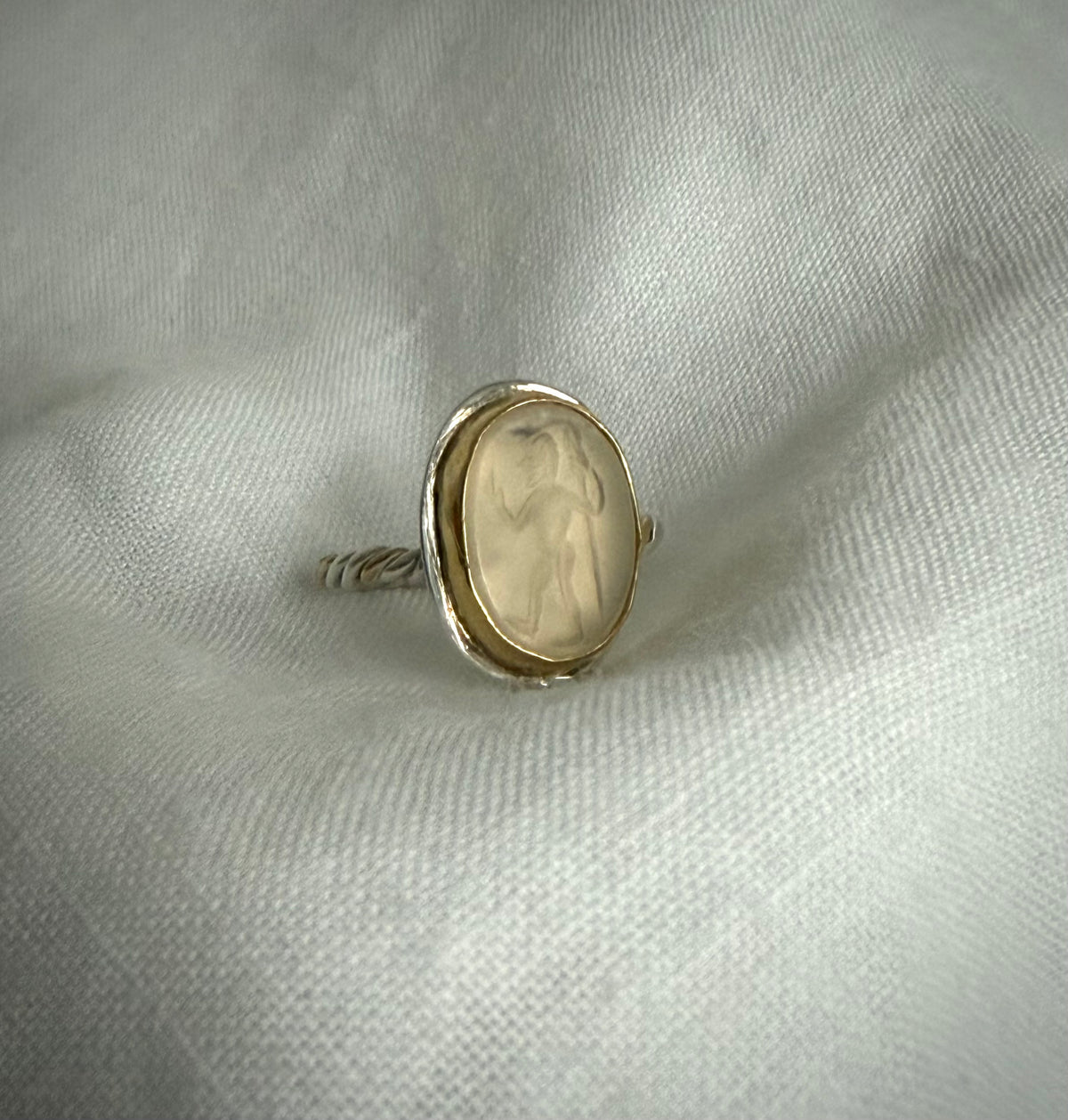 Silver ring with a yellow gold bezel surrounds clear crystal quartz a stone on a textured white fabric background. The stone is carved with a figure walking holding a staff.