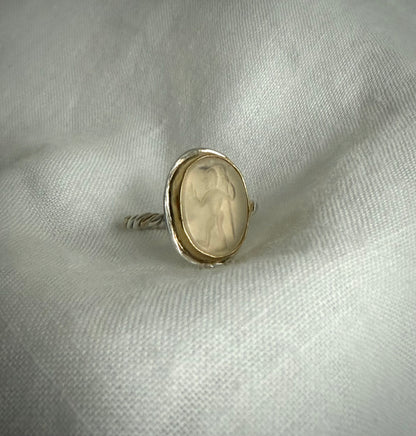 Silver ring with a yellow gold bezel surrounds clear crystal quartz a stone on a textured white fabric background. The stone is carved with a figure walking holding a staff.